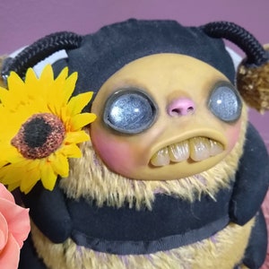 Puede incluir: Un juguete de peluche de abeja con un cuerpo rayado negro y amarillo, antenas marrones y peludas y un gran girasol sujeto a su cabeza. La abeja tiene ojos grandes, redondos y azules y una amplia sonrisa con dientes.