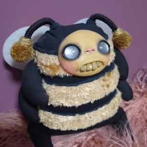 May include: A plush bee toy with a black and yellow striped body, a black head with large white eyes, and a toothy grin. The bee has a fuzzy texture and is sitting on a pink fuzzy surface.