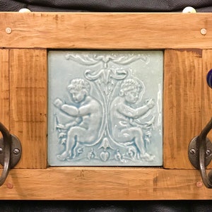May include: Wooden wall-mounted coat rack with a light blue tile featuring cherubs. The rack has two dark metal hooks with blue ceramic knobs. The frame is made of light-colored wood.