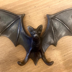 May include: A cast iron bat-shaped wall hook with outstretched wings. The bat is dark grey with a textured finish, featuring a detailed face and wing structure. The hook is designed for wall mounting.