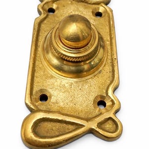 Può includere: Un campanello in ottone vintage con una placca posteriore rettangolare decorativa. Il pulsante ha una parte superiore arrotondata e una base a coste. La placca posteriore presenta disegni curvi ornati nella parte superiore e inferiore e ha tre fori per le viti.