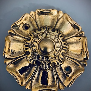Può includere: Campanello in ottone dorato ornato con un motivo floreale. Il campanello presenta un pulsante centrale circondato da dettagli di volute e forme a petalo. L'ottone ha un aspetto lucido e invecchiato, con tre piccoli fori.