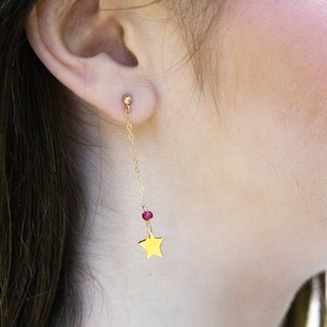 May include: Gold star dangle earrings with a red bead on a gold chain. The earrings are worn by a person with long dark hair.