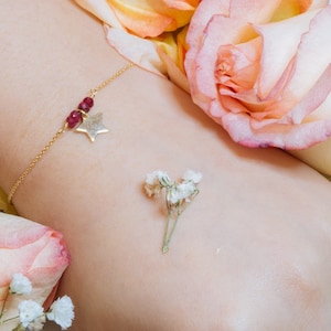 May include: A gold chain bracelet with a star charm and red gemstone accents. The bracelet is on a person's wrist with pink roses and white flowers in the background.