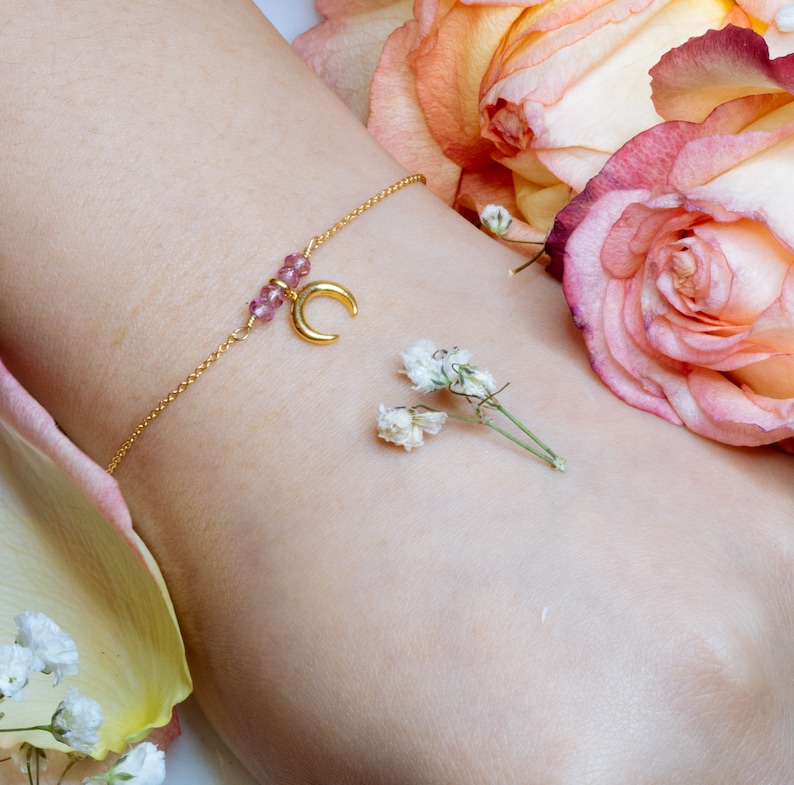 Puede incluir: Un brazalete de cadena dorada con un dije de media luna y cuentas rosadas. El brazalete est&aacute; en la mu&ntilde;eca de una persona y est&aacute; rodeado de flores rosas y blancas.
