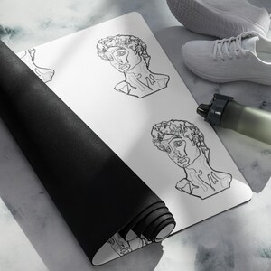 Peut inclure: Tapis de yoga blanc avec un bord roulé noir, orné d'un dessin au trait répété d'un buste classique. Une paire de chaussures de sport blanches et une gourde vert foncé sont également visibles.