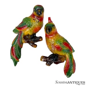 May include: Two colorful ceramic parrot figurines with green, yellow, red, and black paint. The parrots are perched on branches and have their wings spread out.
