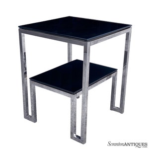 May include: A chrome-plated end table with two black glass shelves. The table has a square top and a rectangular bottom shelf.