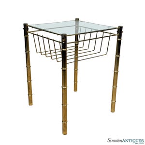 May include: A vintage side table with a clear glass top and a brass-colored metal frame. The table features a magazine rack and bamboo-style legs. The table is square and has a classic, mid-century modern design.