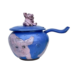 May include: A blue ceramic serving dish with a lid and a handle. The dish is decorated with pink pig illustrations. A small pink pig figurine sits on top of the lid.