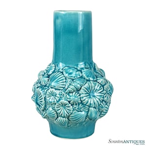 May include: A turquoise ceramic vase with a textured surface. The vase features a cylindrical neck and a bulbous base adorned with seashell designs. The vase is approximately 10 inches tall.