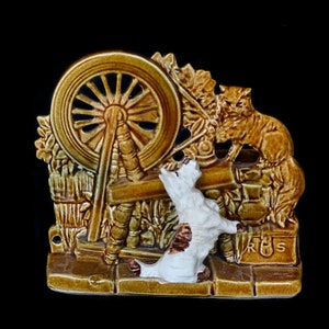 May include: A ceramic wall plaque depicting a dog and a cat by a spinning wheel. The dog is white with brown spots and is looking up at the cat. The cat is brown and is sitting on the spinning wheel. The plaque is decorated with green and brown leaves and has the letters "R.S." on the bottom.