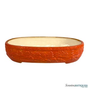May include: Oval-shaped orange ceramic planter with a cream-colored interior. The exterior features a detailed relief of a landscape with buildings and trees. A decorative piece for plants or home decor.