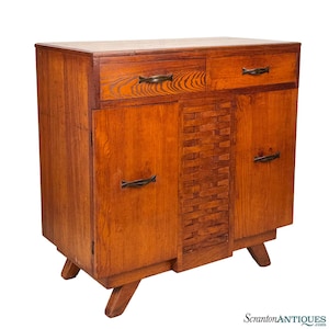 May include: A wooden cabinet with two drawers and two doors. The cabinet is made of a light brown wood with a woven pattern in the center. It has dark metal handles and sits on angled legs.