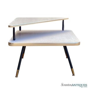 May include: A two-tiered mid-century modern side table with a white laminate top and black metal legs. The top tier is angled and has a gold trim. The legs have gold accents.