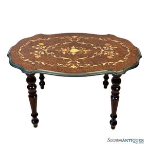 May include: Oval-shaped antique table with a dark brown wood grain top. The table features an inlaid floral design in shades of yellow, orange, and cream. The table has four dark brown, turned legs. The table is a decorative furniture piece.