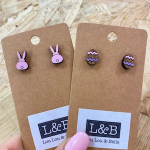 May include: Two sets of wooden earrings on brown card stock. One set features pink bunny rabbit earrings and the other set features purple and brown striped egg earrings. The card stock has a white label with the text "L&B Lou Lou & Belle".