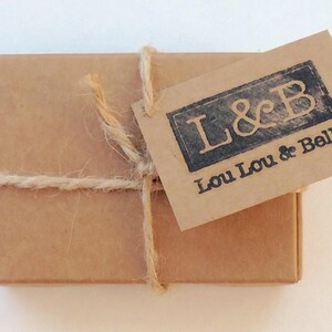 May include: A small, rectangular brown cardboard box tied with twine. A matching brown tag is attached, stamped with the text "L&B" above "Lou Lou & Belle" in a dark, rectangular frame.