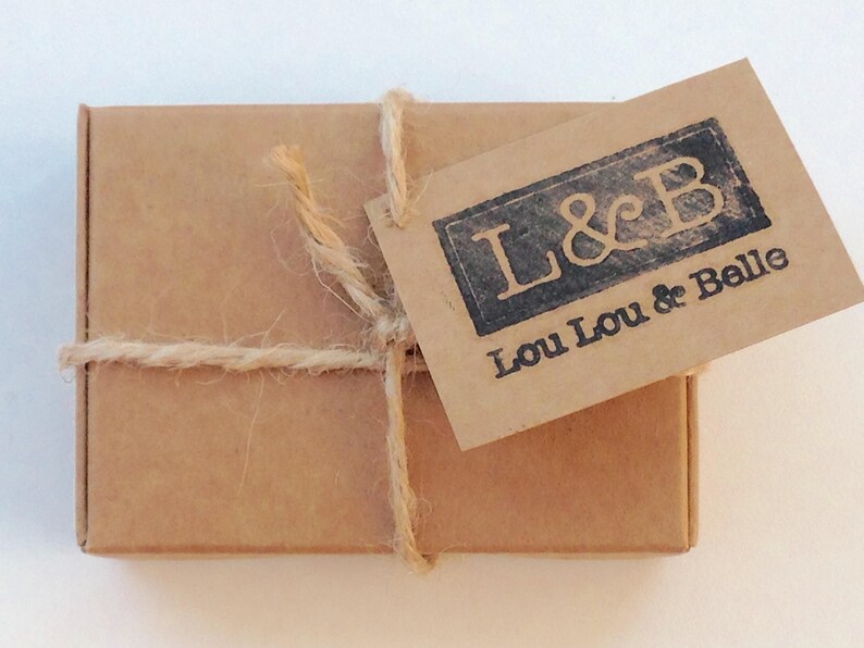May include: A small, rectangular brown cardboard box tied with twine. A matching brown tag is attached, stamped with the text "L&B" above "Lou Lou & Belle" in a dark, rectangular frame.