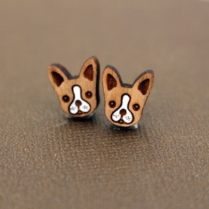 May include: A pair of wooden stud earrings featuring a French Bulldog design. The earrings are light brown with dark brown accents on the ears and a white and black face. The earrings are on a brown surface.