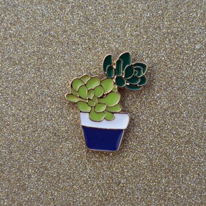Op de afbeelding: Een gouden emaille pin met een groene succulent in een blauw-witte pot.