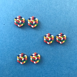 Puede incluir: Cinco pendientes de madera en forma de cubo, cada uno con un patrón diferente de cuadrados rojos, azules, amarillos, verdes y blancos. Los pendientes están dispuestos en forma de escalera en un fondo azul.