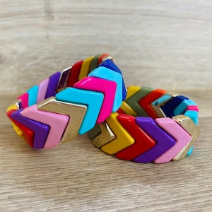 Könnte beinhalten: Zwei bunte Chevron-Muster-Armbänder mit goldenen Akzenten. Die Armbänder sind aus Kunststoff und übereinander gestapelt.
