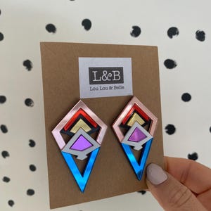 Pode incluir: Brincos geométricos em forma de diamante, com acrílico espelhado rosa, vermelho, dourado e azul. Os brincos estão em um cartão marrom com o texto "L&B Lou Lou & Belle".