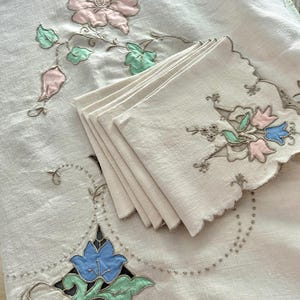 Pode incluir: Toalha de mesa e guardanapos de linho creme. A toalha de mesa apresenta desenhos florais bordados em rosa, azul e verde. Os guardanapos são empilhados e têm bordas onduladas com bordados florais semelhantes.