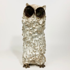 Könnte beinhalten: Weiße Keramik-Eulen-Skulptur mit braunen Augen. Die Eule hat eine strukturierte Oberfläche und steht auf einem weißen Hintergrund.