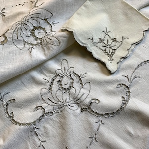 Pode incluir: Toalha de mesa de linho branco com delicados desenhos florais bordados. A toalha de mesa apresenta trabalhos de renda intrincados e uma borda festonada.