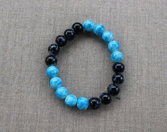 Peut inclure: Un bracelet de perles rondes alternant des perles noires et bleu-vert. Le bracelet élastique est présenté sur une surface grise. Les perles ont un aspect marbré. Circonférence d'environ 19 cm.