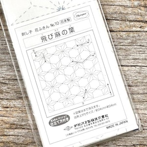 Op de afbeelding: Een Hana-Fukin Sashiko Sampler-kit met Japanse tekst. De kit bevat witte stof met een geometrisch patroon en instructies. De verpakking is blauw en wit, met de merknaam "Olympus". Afmeting afgewerkt: ongeveer 34 cm x 34 cm.