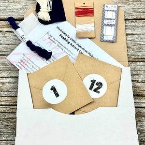 May include: A craft kit featuring a cardboard box containing two small brown envelopes labelled with the numbers 1 and 12, a pack of needles, spools of red and white thread, a bundle of dark blue thread, a white fabric swatch, and instructions.