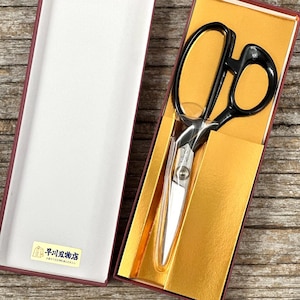 May include: A pair of black-handled scissors with silver blades, presented in a red and gold box. The box is open, revealing the scissors and a white interior. The scissors are likely for sewing or crafting.