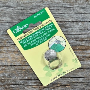 Könnte beinhalten: Ein Clover Ringfingerhut mit Platte, verpackt auf einer gelb-grünen Karte. Der Fingerhut ist silberfarben und hat eine strukturierte Oberfläche. Die Verpackung enthält Text in mehreren Sprachen.