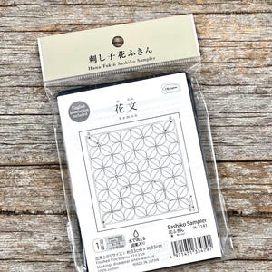 Pode incluir: Um kit Hana-Fukin Sashiko Sampler embalado. O kit inclui tecido azul marinho com um padrão floral, instruções em inglês e o texto "花文" (Kamon). O tamanho final é de aproximadamente 33 cm x 33 cm.