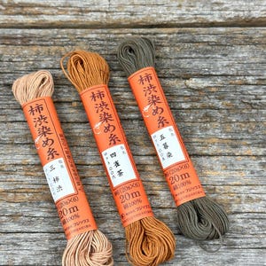 May include: Three spools of thread in shades of brown and green, each wrapped in orange paper with Japanese text. The spools are labeled with the thread type, colour, and length of 20 metres.