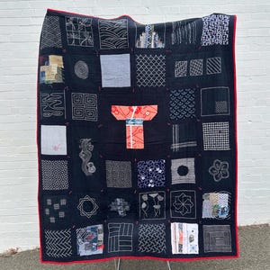 May include: A black quilt with a red border, featuring various square patches with white embroidered designs. A central patch depicts a kimono. The quilt is displayed against a white brick wall.