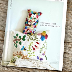 Può includere: Un libro intitolato "Embroidery sewed with the darning stitch" con un orsetto ricamato colorato e un cuscino bianco con ricami floreali. Il libro ha testo giapponese ed è su una superficie di legno.