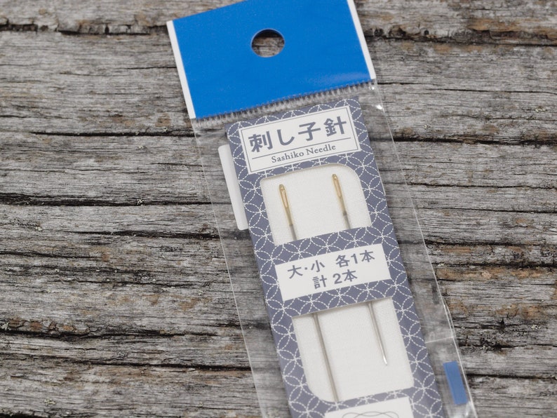 May include: A package of two Sashiko needles, one large and one small. The package is white with blue and gray accents and Japanese text that reads "Sashiko Needle" and "Large: 1 Needle, Small: 2 Needles".