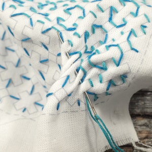 May include: Close-up of a white fabric with blue embroidery. The fabric is folded and a needle with blue thread is visible.