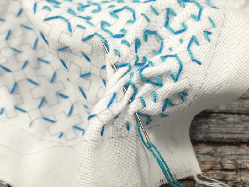 May include: Close-up of a white fabric with blue embroidery. The fabric is folded and a needle with blue thread is visible.
