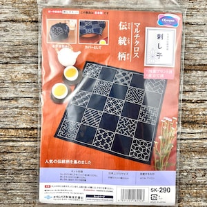 May include: A Sashiko embroidery kit in a clear plastic package featuring a dark blue fabric square with white geometric patterns. The packaging displays the Olympus brand logo, Japanese text, and images showing the finished cloth used as a wrap.