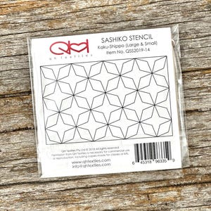 May include: A clear plastic package containing a Sashiko stencil. The stencil features a repeating geometric star pattern. The package includes the text "SASHIKO STENCIL" and "Kaku-Shippo (Large & Small)". The background is a wooden surface.