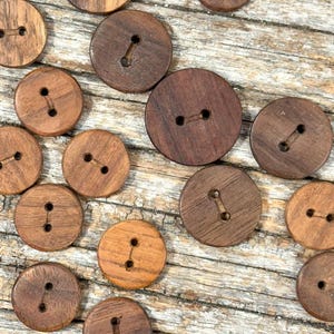May include: A selection of circular wooden buttons in shades of brown. Each button features two sewing holes. The buttons are arranged on a weathered wooden surface, highlighting the natural wood grain.