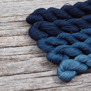 May include: Four skeins of yarn in shades of blue, ranging from dark indigo to light teal, arranged in a gradient on a weathered wooden surface.