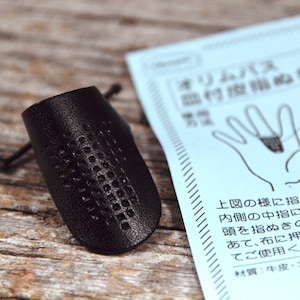 Puede incluir: Un dedal de cuero negro con un patrón de cuadrícula en la superficie. El dedal está diseñado para proteger el dedo mientras se cose. La imagen incluye instrucciones en japonés para usar el dedal.