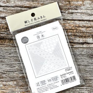 Pode incluir: Um kit de amostra Sashiko embalado com texto japonês e inglês "Hana-Fukin Sashiko Sampler". O kit inclui um tecido branco com vários padrões geométricos para costura e instruções em inglês. O tamanho final é de aproximadamente 34 cm x 34 cm.