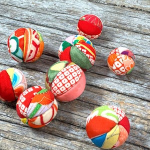 Puede incluir: Una colección de pequeñas bolas de tela coloridas con varios patrones y diseños. Las bolas presentan una mezcla de telas rojas, naranjas, verdes y azules, creando una estética vibrante y juguetona. Están dispuestas sobre una superficie de madera envejecida.